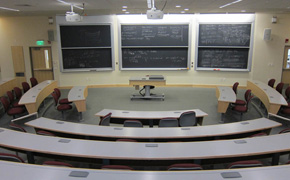 View of the classroom from the very back of the room facing the sliding chalkboards, showing the tiered student desks.