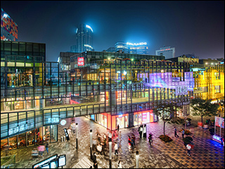 A photograph of a city at night time. There is a plaza in the foreground, a large glass building in the middle, and a city skyline in the background. Many of the buildings have neon lights of all colors. The lights are reflected off the glass building.