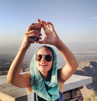 A photograph of a woman holding a cell phone in front of her taking a picture of herself against a landscape of mountains.