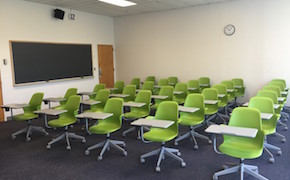A well-lit, medium-sized carpeted classroom with five rows of green moveable furniture tablet armchairs.