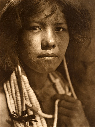 A sepia-toned photograph of a Native American young woman.  She has beaded necklaces wrapped around her neck and is looking directly into the camera.