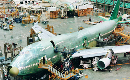 The shell of an airplane being built sits in a giant hangar with lots of equipment surrounding it. 