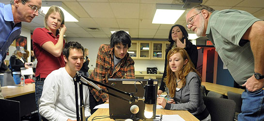 Five students and two educators sit and stand around a table to examine some scientific equipment.