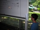 A student kneels by the whiteboard and looks through a hole in a piece of paper taped to the side.