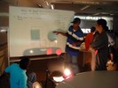 Students stand around a whiteboard with a lit lightbulb casting shadows on the board.