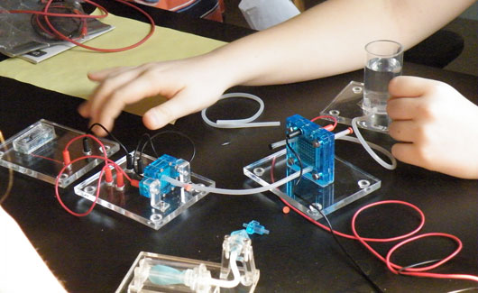 Various wires and circuits sit on a lab bench with students standing around.